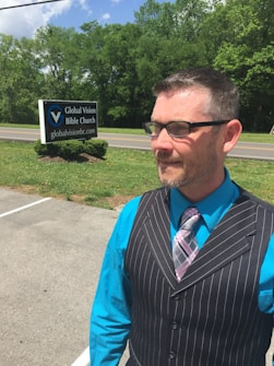 A man wearing a black pinstripe vest over a bright turquoise shirt with a plaid tie stands outdoors near a sign for Global Vision Bible Church. The background features a road and a lush, green forest under a partly cloudy sky.