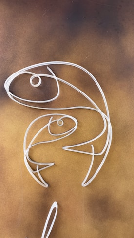 Wire sculptures in the shape of two abstract fish are set against a brown, textured background. The design is minimalist and elegant, with smooth curves and a simple structure.
