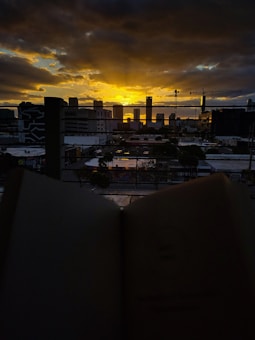 A cityscape at sunset with a dramatic sky filled with dark, looming clouds and streaks of sunlight breaking through near the horizon. In the foreground, blurred pages of an open book create a thematic juxtaposition with the industrial skyline and construction cranes visible in the background.