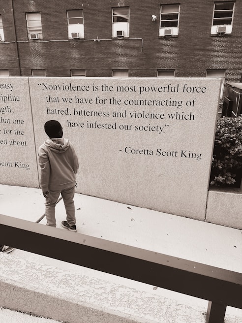 A person standing in front of a wall that features a quote by Coretta Scott King about nonviolence. The background consists of a brick building with several windows, and the scene has a monochromatic color scheme.