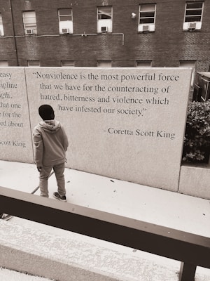 A person standing in front of a wall that features a quote by Coretta Scott King about nonviolence. The background consists of a brick building with several windows, and the scene has a monochromatic color scheme.