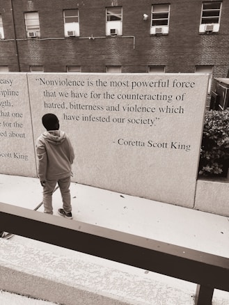 A person standing in front of a wall that features a quote by Coretta Scott King about nonviolence. The background consists of a brick building with several windows, and the scene has a monochromatic color scheme.