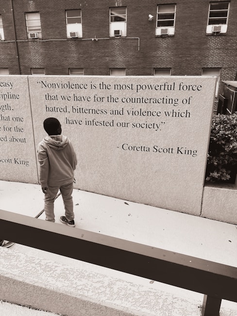 A person standing in front of a wall that features a quote by Coretta Scott King about nonviolence. The background consists of a brick building with several windows, and the scene has a monochromatic color scheme.