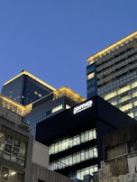Tall modern office buildings with illuminated windows rise against a clear evening sky. The architecture features sleek glass and metal facades, with a prominent sign reading 'pmo' on one of the buildings.