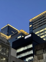 Tall modern office buildings with illuminated windows rise against a clear evening sky. The architecture features sleek glass and metal facades, with a prominent sign reading 'pmo' on one of the buildings.
