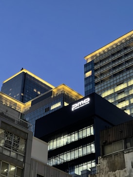 Tall modern office buildings with illuminated windows rise against a clear evening sky. The architecture features sleek glass and metal facades, with a prominent sign reading 'pmo' on one of the buildings.