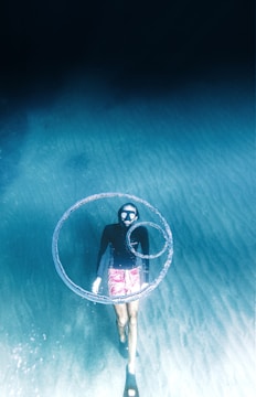 A diver with a snorkel mask swims underwater, releasing swirling air bubbles. The diver wears a dark wetsuit and pink shorts, surrounded by clear blue water with a sandy seabed visible beneath.