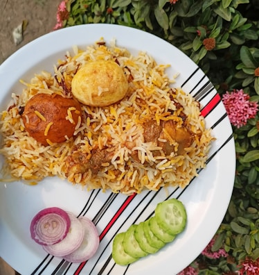 A plate of biryani with pieces of chicken and two boiled eggs is garnished with slices of red onion and cucumber. The dish is placed on a patterned white plate, and it is set against a background of green foliage with some pink flowers.