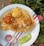 A plate of biryani with pieces of chicken and two boiled eggs is garnished with slices of red onion and cucumber. The dish is placed on a patterned white plate, and it is set against a background of green foliage with some pink flowers.