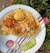 A plate of biryani with pieces of chicken and two boiled eggs is garnished with slices of red onion and cucumber. The dish is placed on a patterned white plate, and it is set against a background of green foliage with some pink flowers.