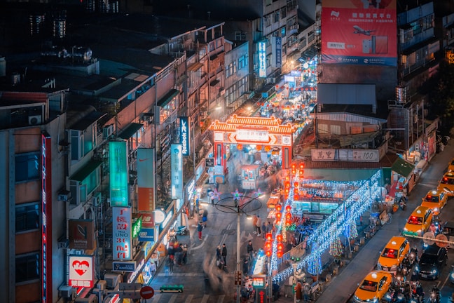 A bustling night market scene in an urban setting. The street is lined with stalls and shops adorned with bright neon signs in various languages. The market entrance is marked by a traditional red and white archway that is illuminated. People are walking along the crowded street, and several yellow taxis are parked alongside.