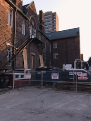 A construction site with a brick building in the background. The building has boarded-up windows and a fire escape. There are construction containers and equipment behind a fence with warning signs.