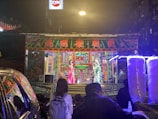 A vibrant street performance taking place on a small stage, adorned with colorful traditional decorations and Chinese characters. Two performers in traditional costumes are on stage, engaging with the audience. Bright lights illuminate the scene, with colorful artwork in the background. Several people are watching the performance from the street. Cotton candy machines are visible in the foreground.