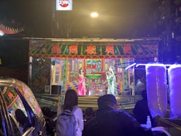 A vibrant street performance taking place on a small stage, adorned with colorful traditional decorations and Chinese characters. Two performers in traditional costumes are on stage, engaging with the audience. Bright lights illuminate the scene, with colorful artwork in the background. Several people are watching the performance from the street. Cotton candy machines are visible in the foreground.