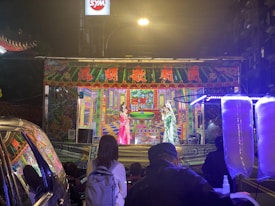 A vibrant street performance taking place on a small stage, adorned with colorful traditional decorations and Chinese characters. Two performers in traditional costumes are on stage, engaging with the audience. Bright lights illuminate the scene, with colorful artwork in the background. Several people are watching the performance from the street. Cotton candy machines are visible in the foreground.