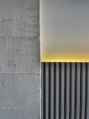 A minimalistic architectural detail featuring a textured concrete wall on the left and a smooth, vertically striped panel on the right. A soft yellow light emanates from the top edge, casting a warm glow on the panel and creating a contrast against the gray tones.