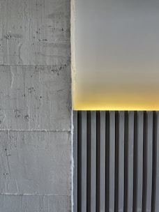 A minimalistic architectural detail featuring a textured concrete wall on the left and a smooth, vertically striped panel on the right. A soft yellow light emanates from the top edge, casting a warm glow on the panel and creating a contrast against the gray tones.