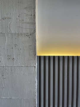 A minimalistic architectural detail featuring a textured concrete wall on the left and a smooth, vertically striped panel on the right. A soft yellow light emanates from the top edge, casting a warm glow on the panel and creating a contrast against the gray tones.