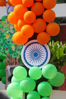 Clusters of orange and green balloons are arranged on a vertical structure. A paper circle featuring the Ashoka Chakra, an element of the Indian national flag, is visible amidst the balloons. Some green leaves are in the background.