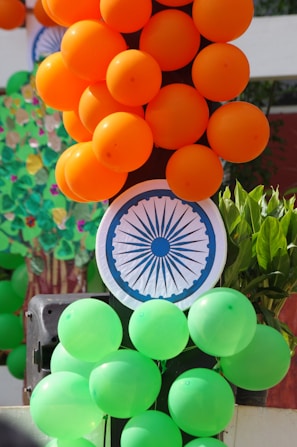 Clusters of orange and green balloons are arranged on a vertical structure. A paper circle featuring the Ashoka Chakra, an element of the Indian national flag, is visible amidst the balloons. Some green leaves are in the background.