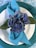 A blue napkin is elegantly folded and placed on a white plate. A blue and purple flower rests on top of the napkin, with green leaves adding contrast. The setting is further enhanced by a turquoise woven placemat beneath the plate and a patterned blue tablecloth in the background.
