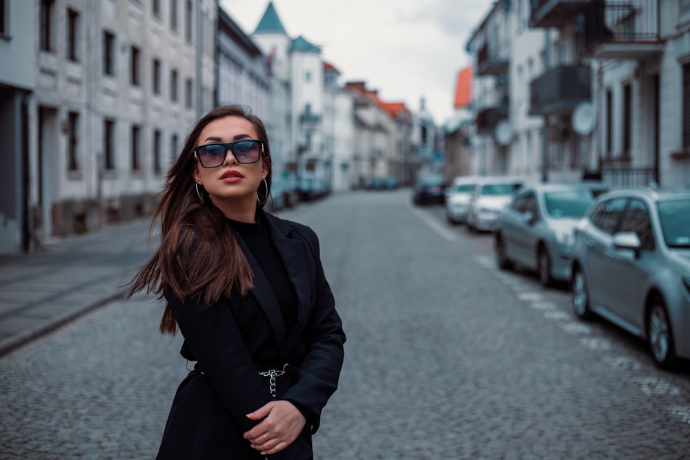 a woman in a black coat and sunglasses standing in the middle of a street