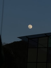 A bright full moon stands out against a clear evening sky. Below, the silhouette of a hill can be seen with a portion of a building on the right, featuring a metal roof and large windows.