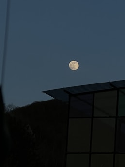A bright full moon stands out against a clear evening sky. Below, the silhouette of a hill can be seen with a portion of a building on the right, featuring a metal roof and large windows.
