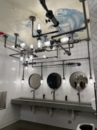 A bathroom with a unique industrial design features exposed piping and cylindrical lights on the ceiling. The ceiling displays a large fish illustration with labeled parts. The walls are covered with white herringbone tiles, and there are three circular mirrors above a long concrete sink with modern faucets.