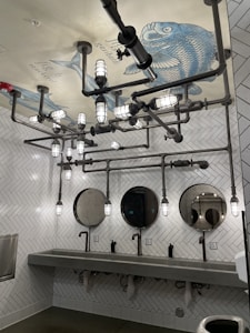 A bathroom with a unique industrial design features exposed piping and cylindrical lights on the ceiling. The ceiling displays a large fish illustration with labeled parts. The walls are covered with white herringbone tiles, and there are three circular mirrors above a long concrete sink with modern faucets.
