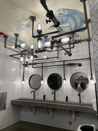 A bathroom with a unique industrial design features exposed piping and cylindrical lights on the ceiling. The ceiling displays a large fish illustration with labeled parts. The walls are covered with white herringbone tiles, and there are three circular mirrors above a long concrete sink with modern faucets.