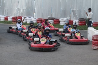 A group of people wearing helmets are seated in go-karts on a race track. Each kart is numbered and has a red color scheme. The track is lined with red and white safety barriers made of tires. One person in the background is standing and looking at a smartphone.