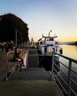 A dockside setting with a large white and blue boat moored by the river. Several people are gathered on the dock, some standing and others sitting, enjoying the evening atmosphere. The sky appears to be at dusk, with gradients of soft blues and warm hues. To the left, a row of trees lines the path alongside the dock.
