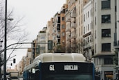 A city street features a bus with route number 61 heading toward Moncloa. Tall residential buildings with various architectural styles line both sides of the street. The trees are bare, suggesting a winter or late autumn setting. Traffic lights and street lamps are visible, and the atmosphere feels overcast and slightly moody.