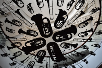A circular ceiling installation features a variety of musical instrument silhouettes, including tubas, trumpets, and trombones. The instruments are suspended uniformly, creating an artistic and dynamic visual effect. The background includes ornate architectural details, such as carved molding and skylight structures.