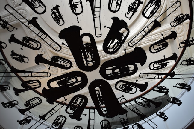 A circular ceiling installation features a variety of musical instrument silhouettes, including tubas, trumpets, and trombones. The instruments are suspended uniformly, creating an artistic and dynamic visual effect. The background includes ornate architectural details, such as carved molding and skylight structures.