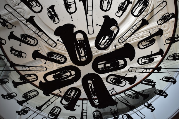 A circular ceiling installation features a variety of musical instrument silhouettes, including tubas, trumpets, and trombones. The instruments are suspended uniformly, creating an artistic and dynamic visual effect. The background includes ornate architectural details, such as carved molding and skylight structures.