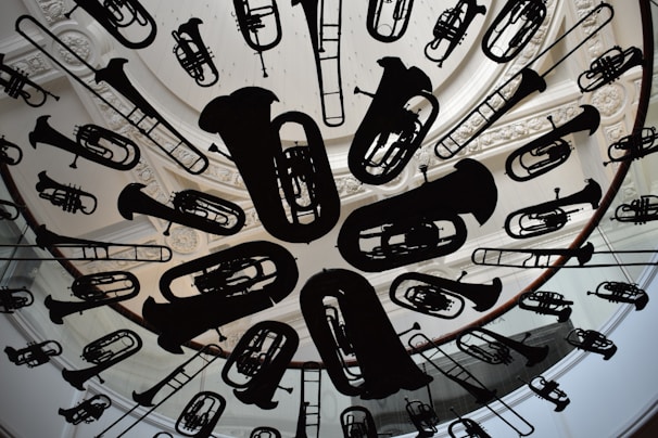 A circular ceiling installation features a variety of musical instrument silhouettes, including tubas, trumpets, and trombones. The instruments are suspended uniformly, creating an artistic and dynamic visual effect. The background includes ornate architectural details, such as carved molding and skylight structures.