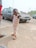 A young child wearing a striped tank top and patterned pants is standing on a pavement. The child is holding earphones, and there is a parked car in the background. Part of an adult wearing red shorts is slightly visible on the left side of the image.