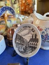 A collection of ceramic items is displayed, including a small decorative plate featuring an illustration of a historic town labeled 'Goslar'. The plate depicts a detailed street scene with traditional architecture, people walking, and a fountain. The background includes various teapots and pitchers with floral and abstract designs.