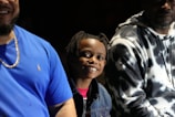 Several people are sitting together with smiles. A child in the center is wearing a denim jacket and has braided hair, looking directly at the camera. Two adults are sitting on either side, one wearing a blue shirt and the other a tie-dye hoodie.