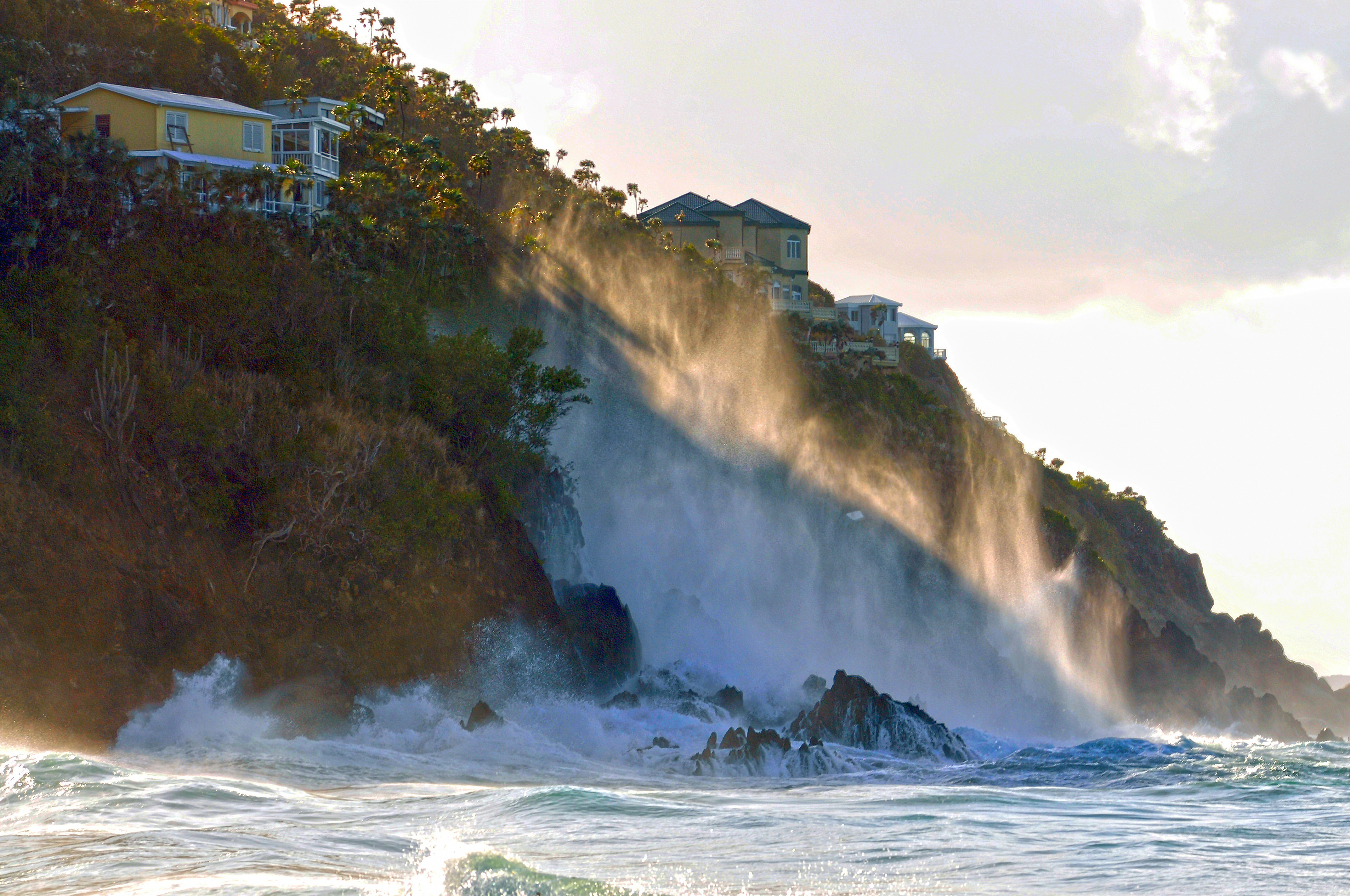 High waves splash and ocean spray flies high up an oceanside cliff side.