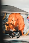 A large mural of a person singing into a microphone is painted on the side of a building. The artwork features vibrant colors, particularly a striking orange used for the hair, complemented by floral patterns in the background. Below the mural, parked cars, including a black SUV, are visible.