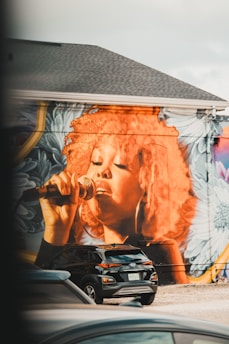 A large mural of a person singing into a microphone is painted on the side of a building. The artwork features vibrant colors, particularly a striking orange used for the hair, complemented by floral patterns in the background. Below the mural, parked cars, including a black SUV, are visible.
