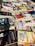 A collection of various books and magazines spread out on a table, with different sizes, colors, and cover designs. The selection includes novels, educational books, and periodicals, some with Spanish titles. The arrangement seems casual, possibly indicating a market or a book fair setting.