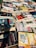 A collection of various books and magazines spread out on a table, with different sizes, colors, and cover designs. The selection includes novels, educational books, and periodicals, some with Spanish titles. The arrangement seems casual, possibly indicating a market or a book fair setting.