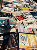 A collection of various books and magazines spread out on a table, with different sizes, colors, and cover designs. The selection includes novels, educational books, and periodicals, some with Spanish titles. The arrangement seems casual, possibly indicating a market or a book fair setting.