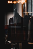 A dark-colored suit jacket is displayed on a mannequin, which is placed in a dimly lit room. Above the jacket, warm golden lights create a cozy ambiance, and reflections on the glass suggest it's inside a shop window.