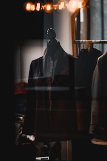 A dark-colored suit jacket is displayed on a mannequin, which is placed in a dimly lit room. Above the jacket, warm golden lights create a cozy ambiance, and reflections on the glass suggest it's inside a shop window.