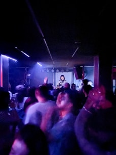 A lively nightclub scene with a crowd of people dancing and enjoying themselves under dim lighting. A DJ is positioned on a raised platform in the background, surrounded by equipment and bright spotlights casting vibrant colors.